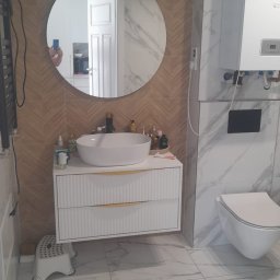 Interfam Tomasz Abramowicz - Nowoczesna łazienka z okrągłym lustrem, białą umywalką na szafce z frezowanymi frontami i marmurowymi płytkami. Widoczny piec gazowy i toaleta.