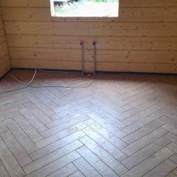 Interfam Tomasz Abramowicz - Ukończona podłoga z płytek ułożonych w jodełkę w drewnianym domku. Widoczne rury instalacji i okno. Niebieska taśma zabezpieczająca przy ścianach.