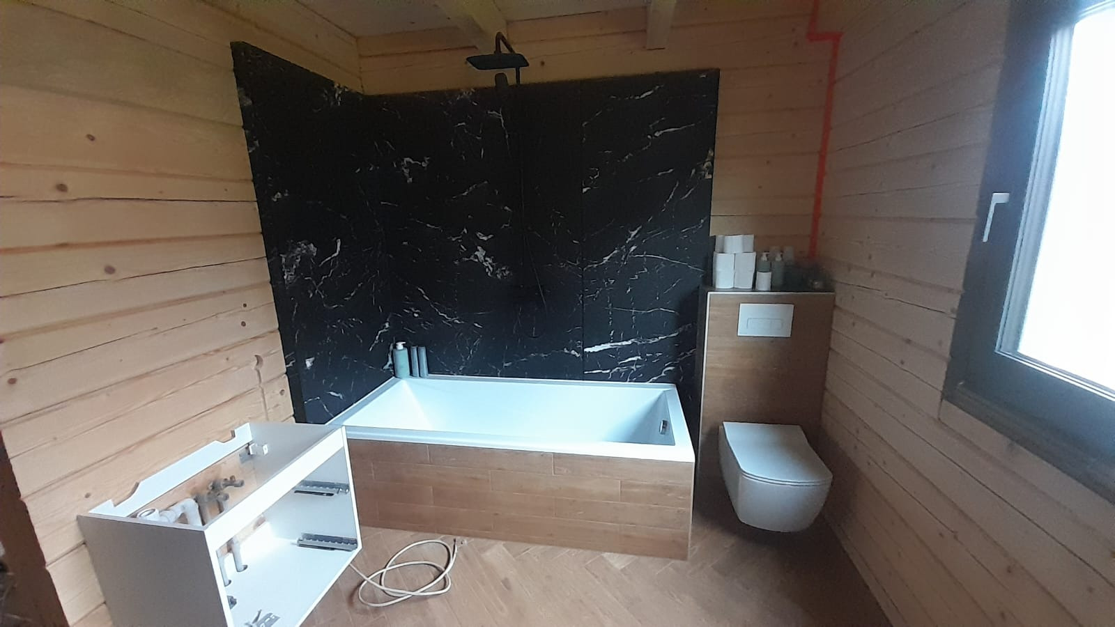 Łazienka w drewnianym domu z czarną ścianą imitującą marmur, białą wanną obudowaną drewnem i wiszącą toaletą. Umywalka w trakcie montażu. Widok na okno.