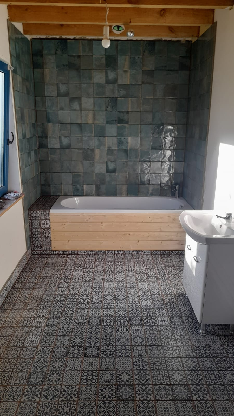 Wykończona łazienka z wanną obudowaną drewnem, umywalką i wzorzystymi płytkami podłogowymi oraz zielonymi płytkami na ścianach. Widoczne drewniane belki stropowe.