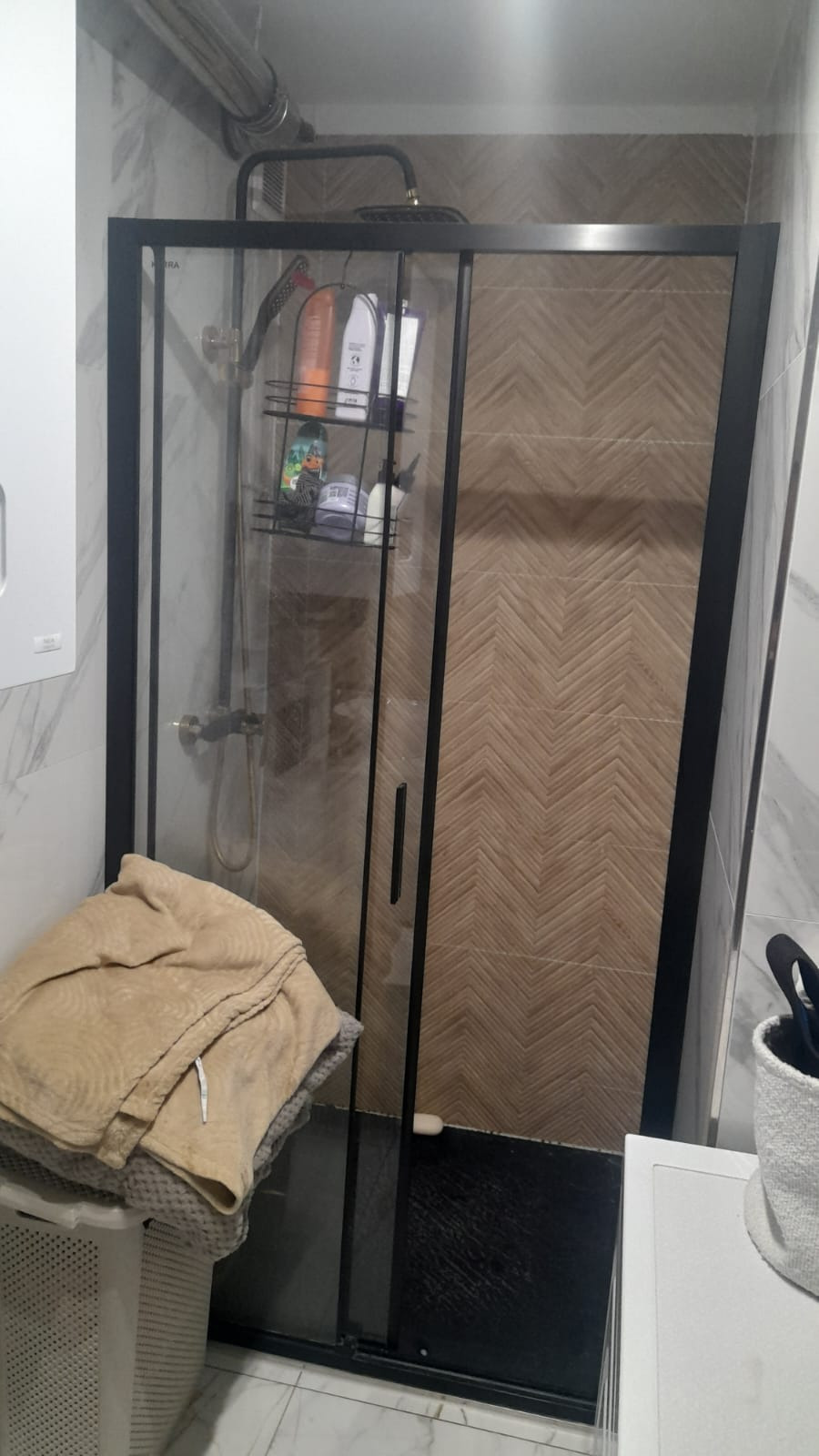 Kabina prysznicowa z czarną ramą i wzorzystymi płytkami w jodełkę na ścianie. Na półce w kabinie widoczne kosmetyki. Obok kosz z ręcznikami.