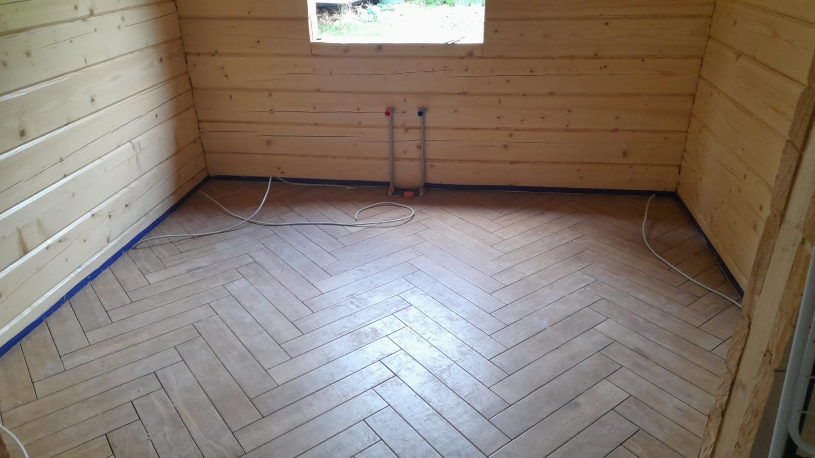 Ukończona podłoga z płytek ułożonych w jodełkę w drewnianym domku. Widoczne rury instalacji i okno. Niebieska taśma zabezpieczająca przy ścianach.