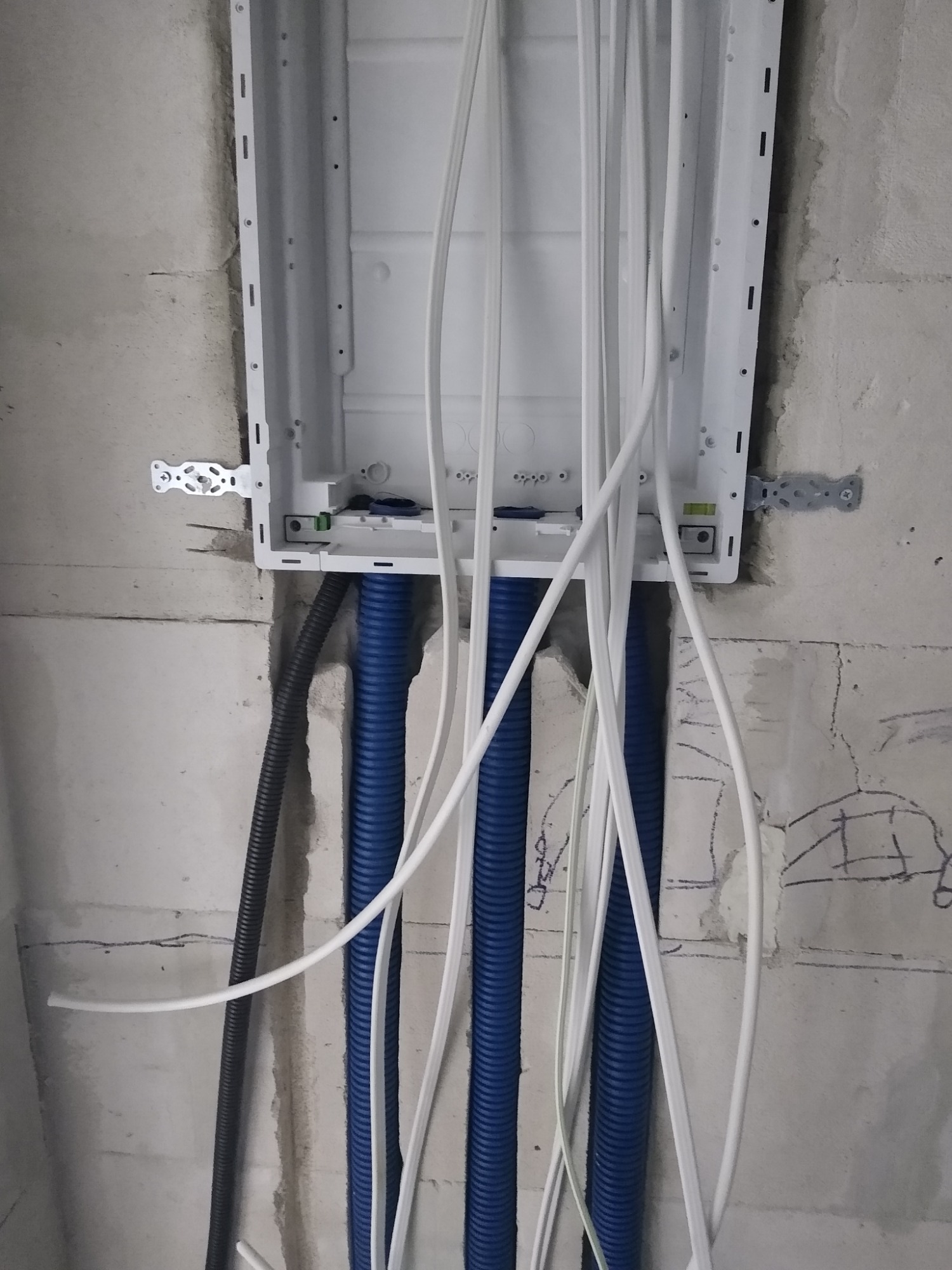 Biała skrzynka elektryczna z poziomnicą zamontowana na surowej ścianie, z wychodzącymi z niej przewodami w białej i niebieskiej karbowanej osłonie, oraz pojedynczym czarnym przewodem karbowanym po...