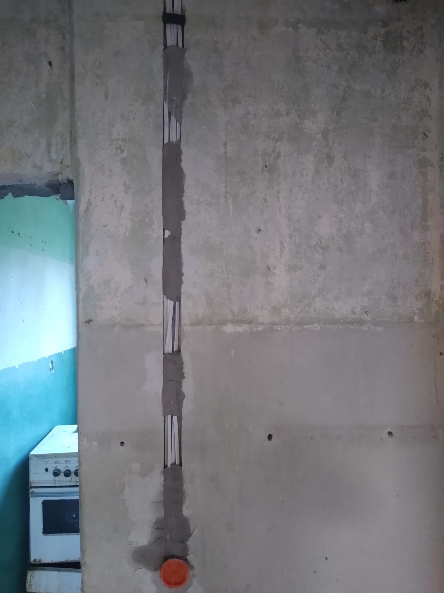 Wewnętrzna instalacja elektryczna w bruździe ściany, z widocznymi przewodami zabezpieczonymi obejmami, częściowo zamaskowana szarym tynkiem, z pomarańczową puszką instalacyjną u dołu, na tle...