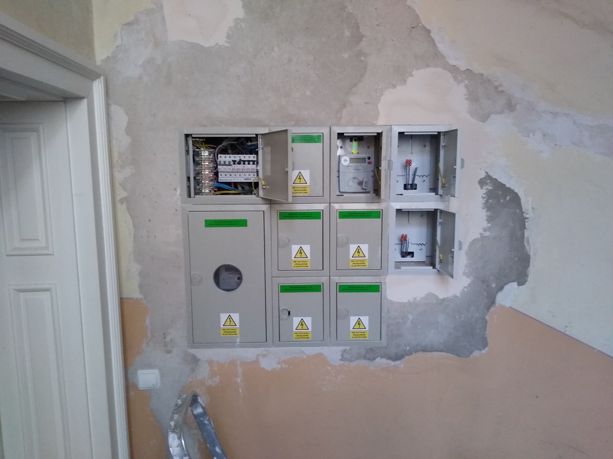 Szafka elektryczna z otwartymi drzwiczkami, widoczne zabezpieczenia i okablowanie, zamontowana na ścianie z widocznymi śladami po remoncie, obok framuga białych drzwi i fragment drabiny.