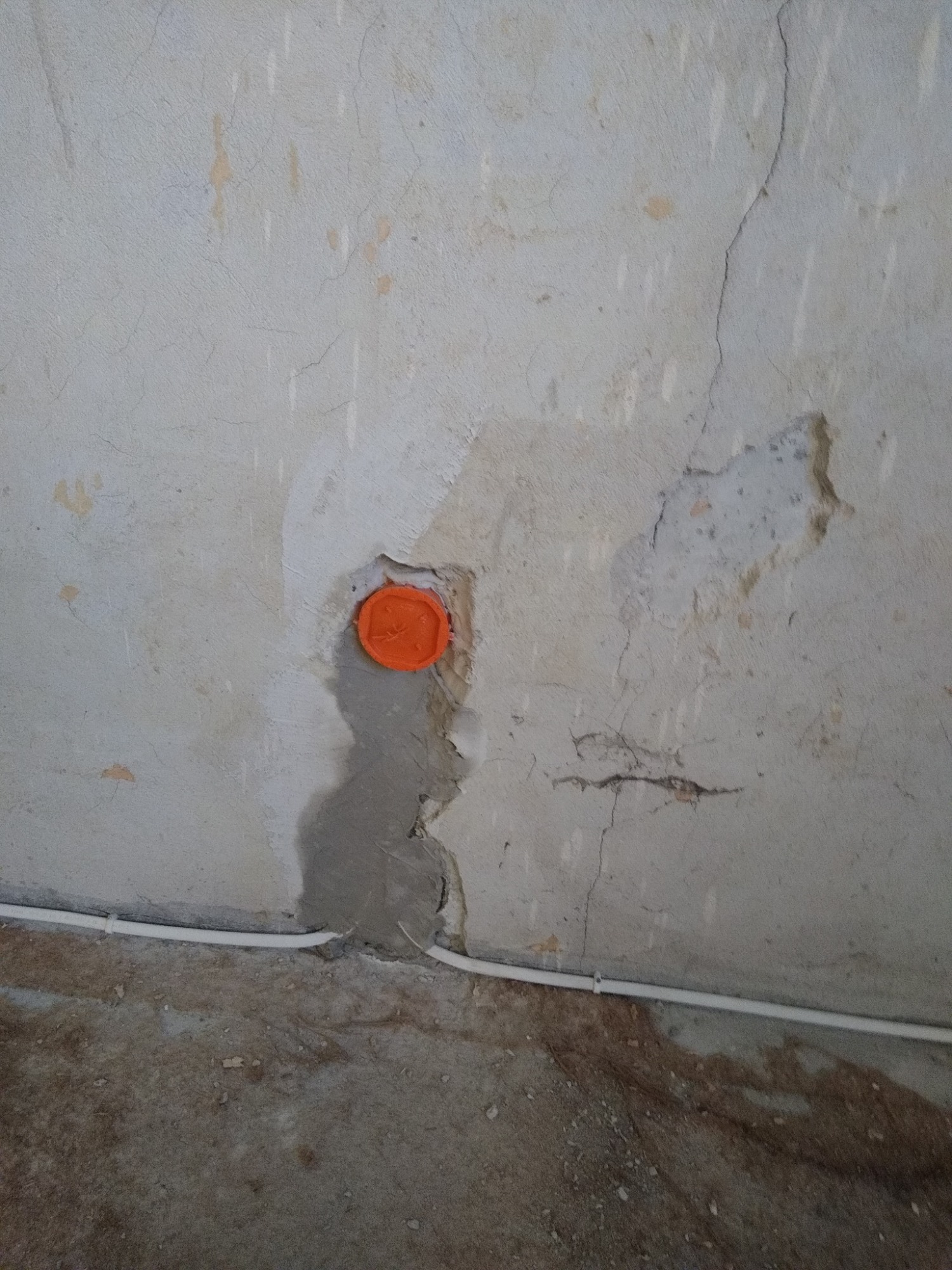 Nierówna ściana z widocznymi pęknięciami i łatami, z pomarańczową puszką instalacyjną wmurowaną na wysokości wzroku i białym kablem elektrycznym biegnącym wzdłuż dolnej krawędzi ściany.
