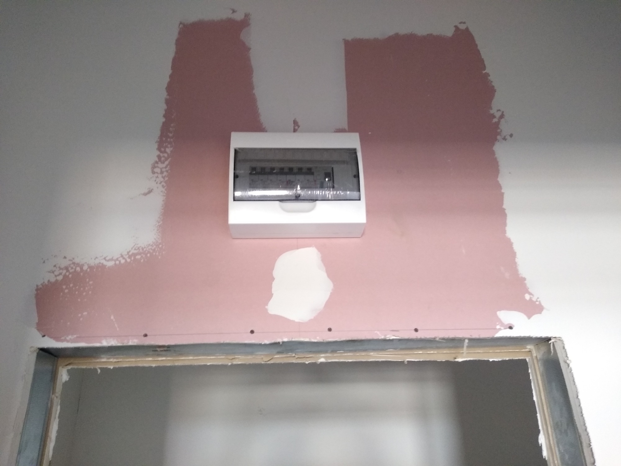 Zamontowana skrzynka elektryczna z przezroczystą osłoną na ścianie z widocznymi śladami po szpachlowaniu i malowaniu, nad surową ościeżnicą drzwiową.