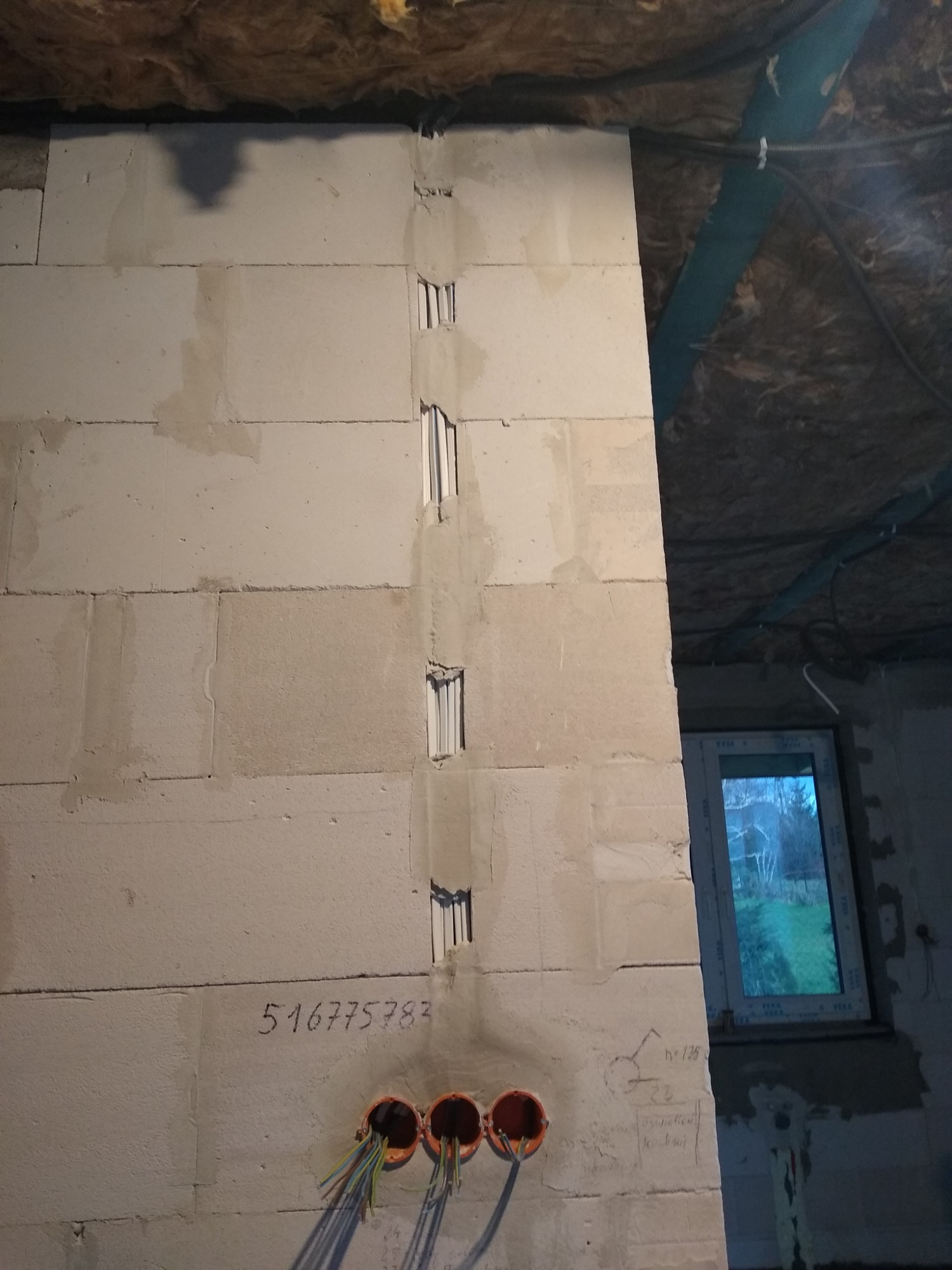 Ściana z betonu komórkowego z wyfrezowanymi pionowymi kanałami pod przewody elektryczne i puszkami instalacyjnymi, widoczne wystające kable, numer telefonu napisany na ścianie, w tle okno...