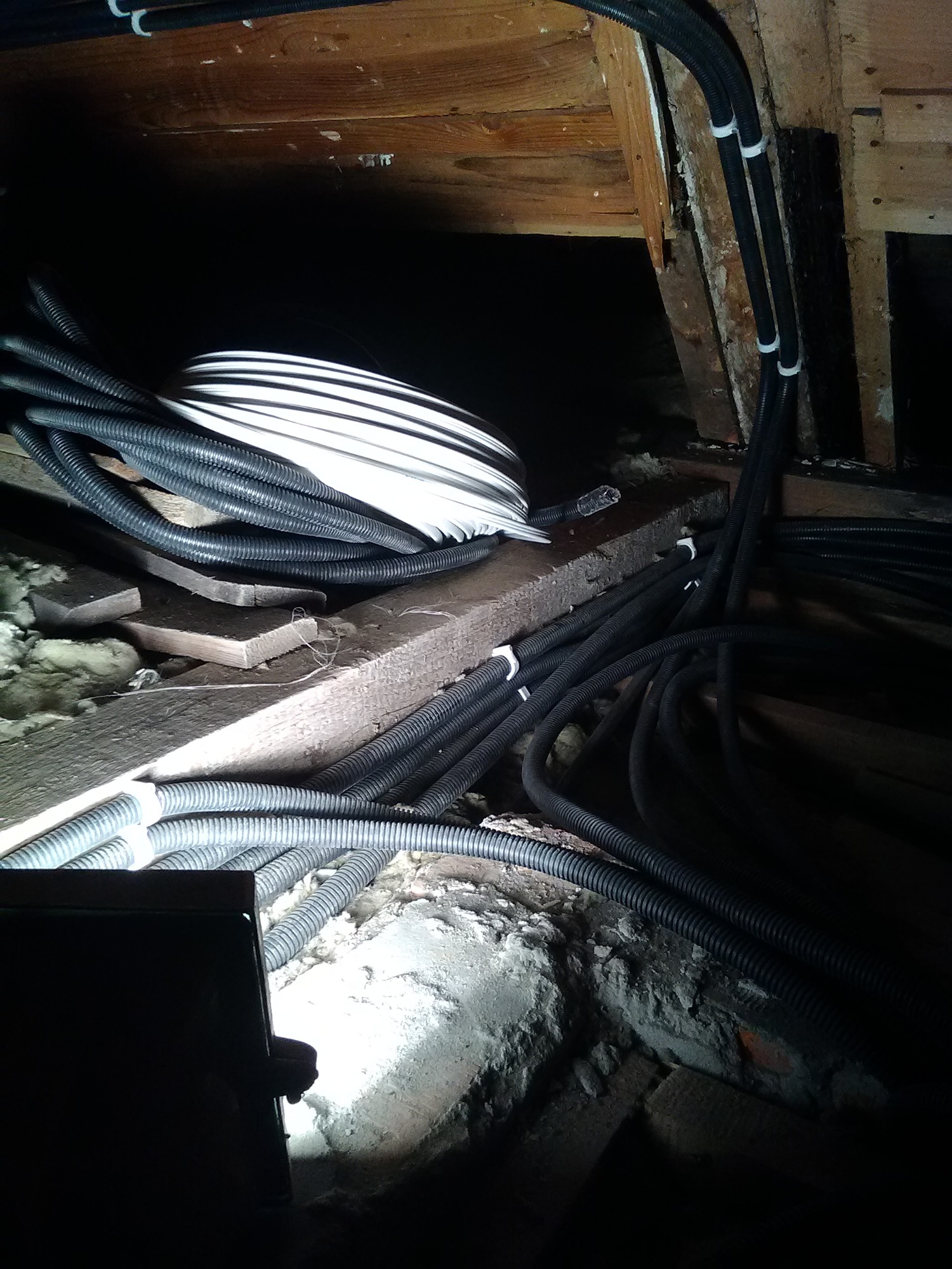 Czarny karbowany peszel z przewodami w przestrzeni poddasza, mocowany białymi uchwytami do drewnianej konstrukcji, widoczne ocieplenie z wełny mineralnej.