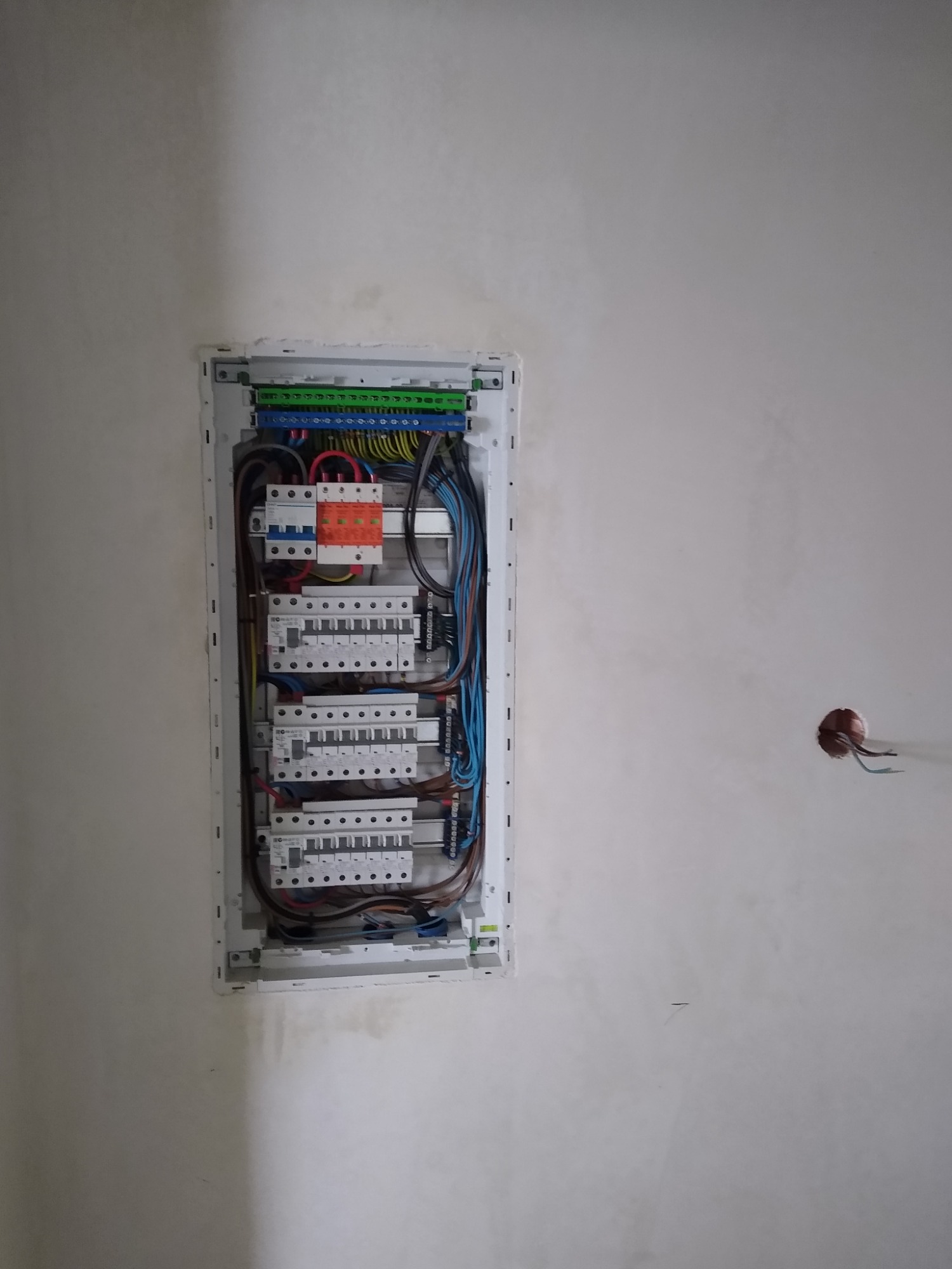 Otwarta skrzynka bezpiecznikowa z widocznymi przewodami, bezpiecznikami i elementami montażowymi na surowej, szarej ścianie z widocznym otworem na gniazdko elektryczne.