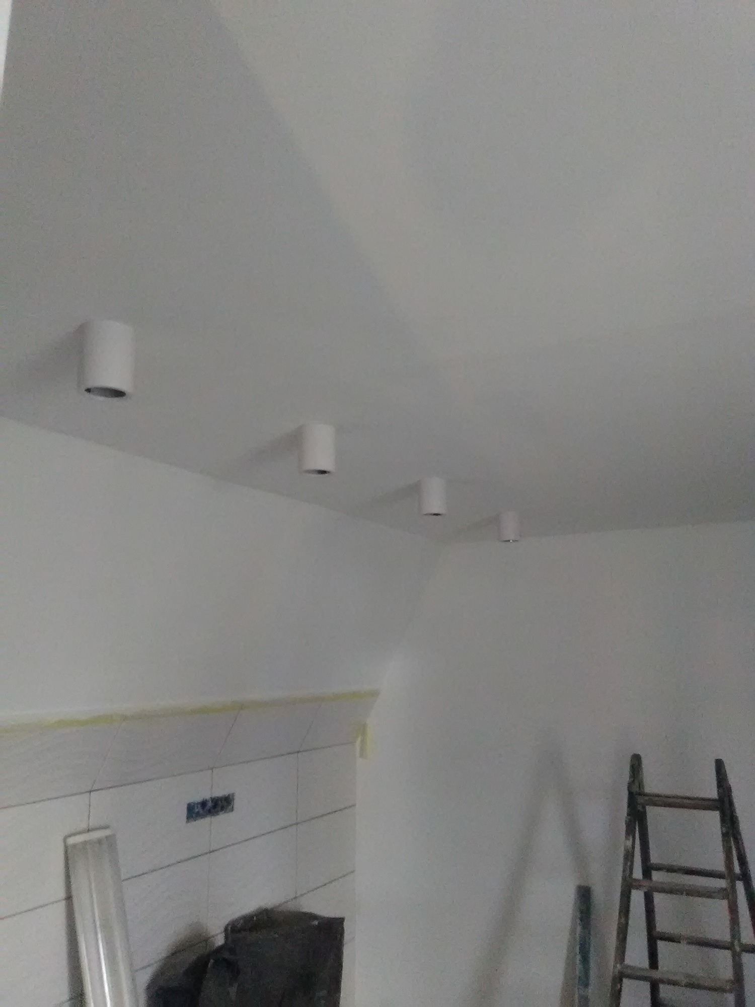 Cztery białe, cylindryczne lampy sufitowe zamontowane w białym pomieszczeniu z widoczną drabina malarską w rogu i częściowo wykończoną ścianą z białymi płytkami.