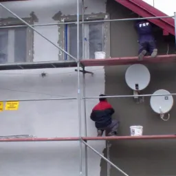 Tynkowanie elewacji domu jednorodzinnego z rusztowania; widoczne anteny satelitarne, okna zabezpieczone folią, tabliczki ostrzegawcze. Częściowo otynkowana ściana.