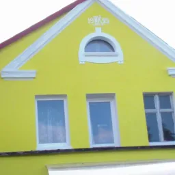 Żółta fasada domu z białymi detalami architektonicznymi, trzema oknami i anteną satelitarną. Widoczny komin i fragment dachu sąsiedniego budynku.