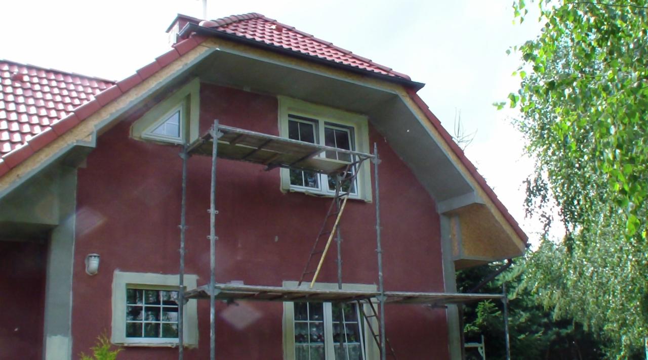 Dom w trakcie prac elewacyjnych: czerwona fasada, okna z białymi ramami, rusztowanie. Widoczne elementy wykończeniowe dachu i obróbki blacharskie. Częściowo otynkowany fragment.