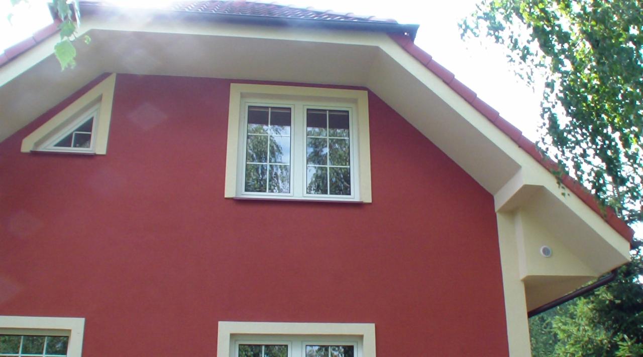 Czerwona elewacja domu z białymi oknami i kremowymi elementami architektonicznymi. Widoczne okno trójkątne i standardowe z refleksami drzew.