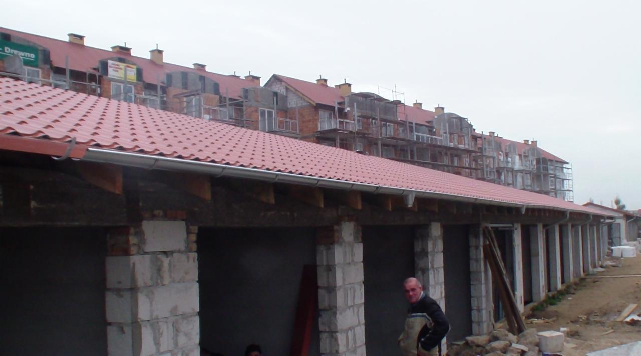 Budynek w trakcie budowy z czerwonym dachem. Widoczne pustaki i fragmenty rusztowań. W tle budynek wielorodzinny w stanie surowym.