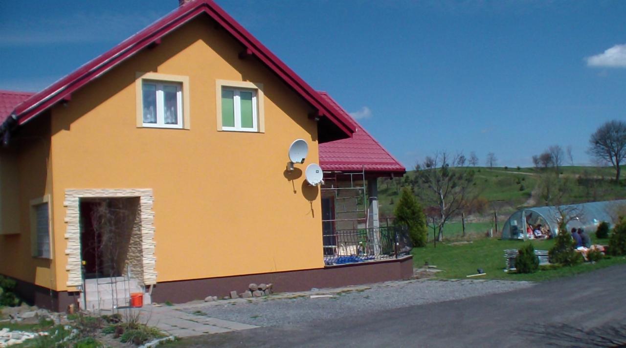Żółty dom z czerwonym dachem i dwiema antenami satelitarnymi w Gardei. Wejście z kamienną obudową. W tle zielony krajobraz i szklarnia.