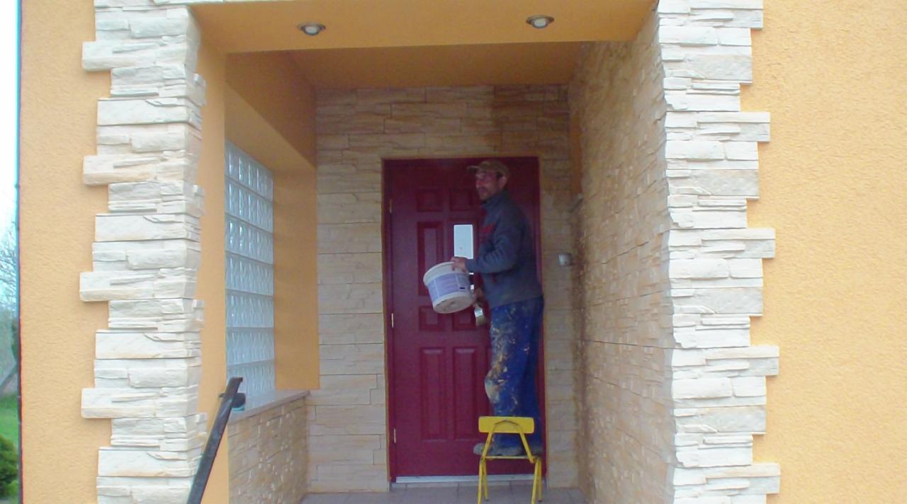 Malarz na żółtym taborecie maluje ścianę przy wejściu do domu na kolor żółty. Dekoracyjne obłożenie ścian imitujące kamień. Czerwone drzwi wejściowe.