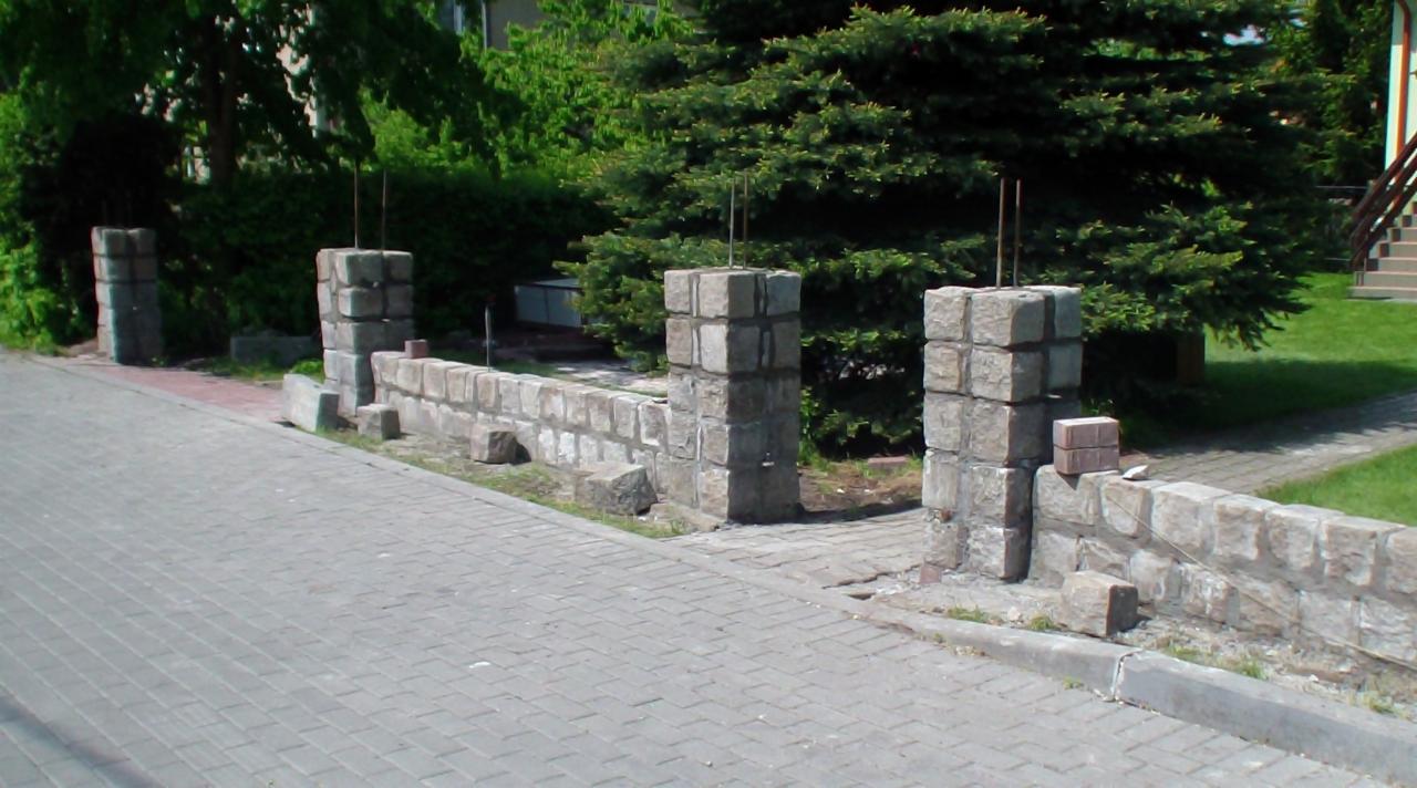 Wznoszenie kamiennego murku z filarami. Widoczne pręty zbrojeniowe wystające z filarów. Częściowo wykonane, surowe wykończenie. Praca w toku.