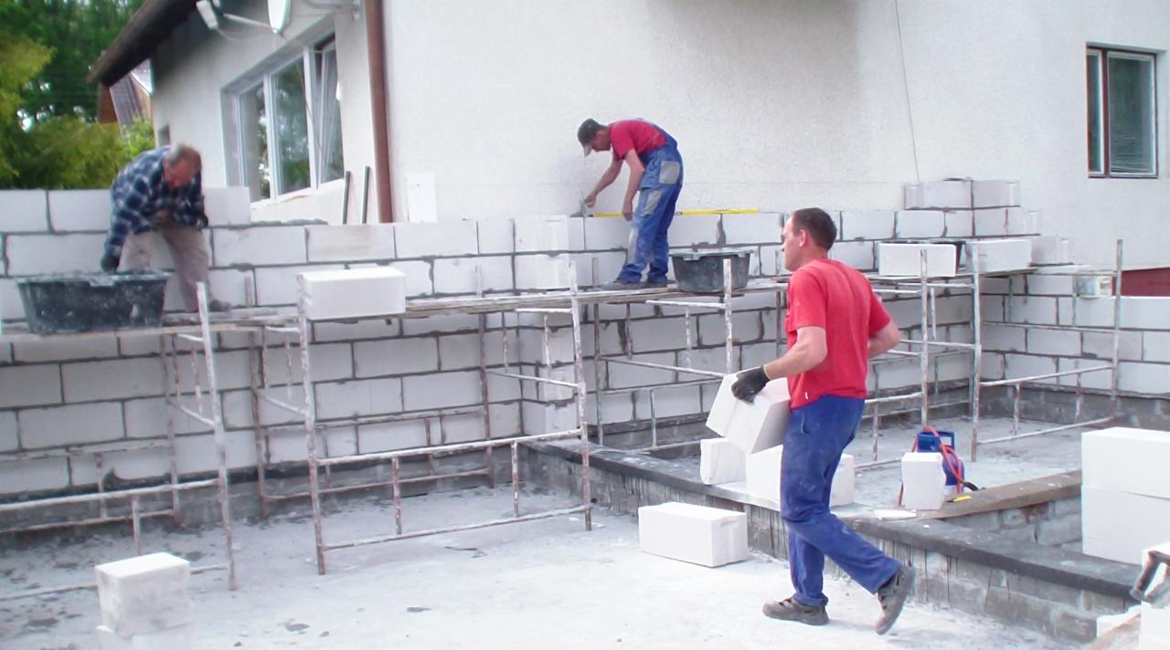 Trzech murarzy w trakcie wznoszenia ściany z białych bloczków na rusztowaniu. Jeden nakłada zaprawę, drugi układa bloczki, trzeci je donosi. Widoczne narzędzia i materiały budowlane.