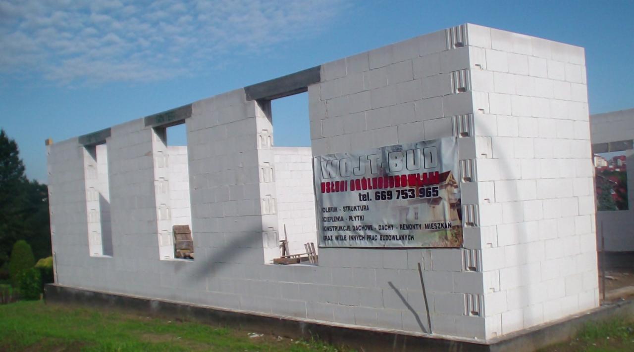 Surowy stan budynku z białych bloczków, widoczne okna i banner reklamowy firmy budowlanej. Fundamenty i początek ścian na tle zieleni i błękitnego nieba.