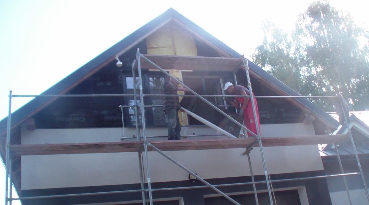 Prace na rusztowaniu przy ocieplaniu poddasza domu jednorodzinnego. Dwóch mężczyzn w trakcie montażu izolacji i folii. Widok z dołu na fragment elewacji i dach.