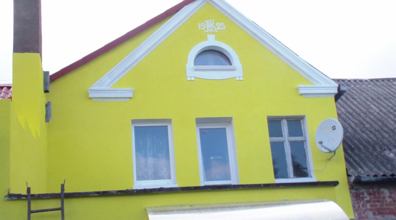 Żółta fasada domu z białymi detalami architektonicznymi, trzema oknami i anteną satelitarną. Widoczny komin i fragment dachu sąsiedniego budynku.