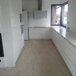 Stolarnia Solarscy - Biała kuchnia na wymiar z połyskiem, widok na okno i sprzęty AGD. Minimalistyczny design, jasne drewnopodobne panele podłogowe, czarny wkład kominkowy po lewej.