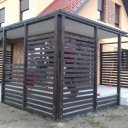 Nowoczesna, ciemnobrązowa konstrukcja z drewnianych listew tworząca zadaszenie tarasu przy domu jednorodzinnym. Widoczne trzy stopnie wejściowe z jasnych płytek oraz fragmenty osób wewnątrz.