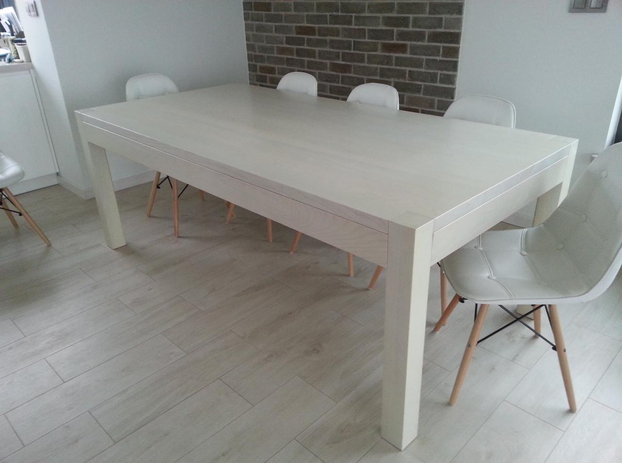 Jasny, prostokątny stół z drewna z szufladą, otoczony białymi krzesłami na drewnianych nogach, na tle ściany z cegły w nowoczesnej jadalni.