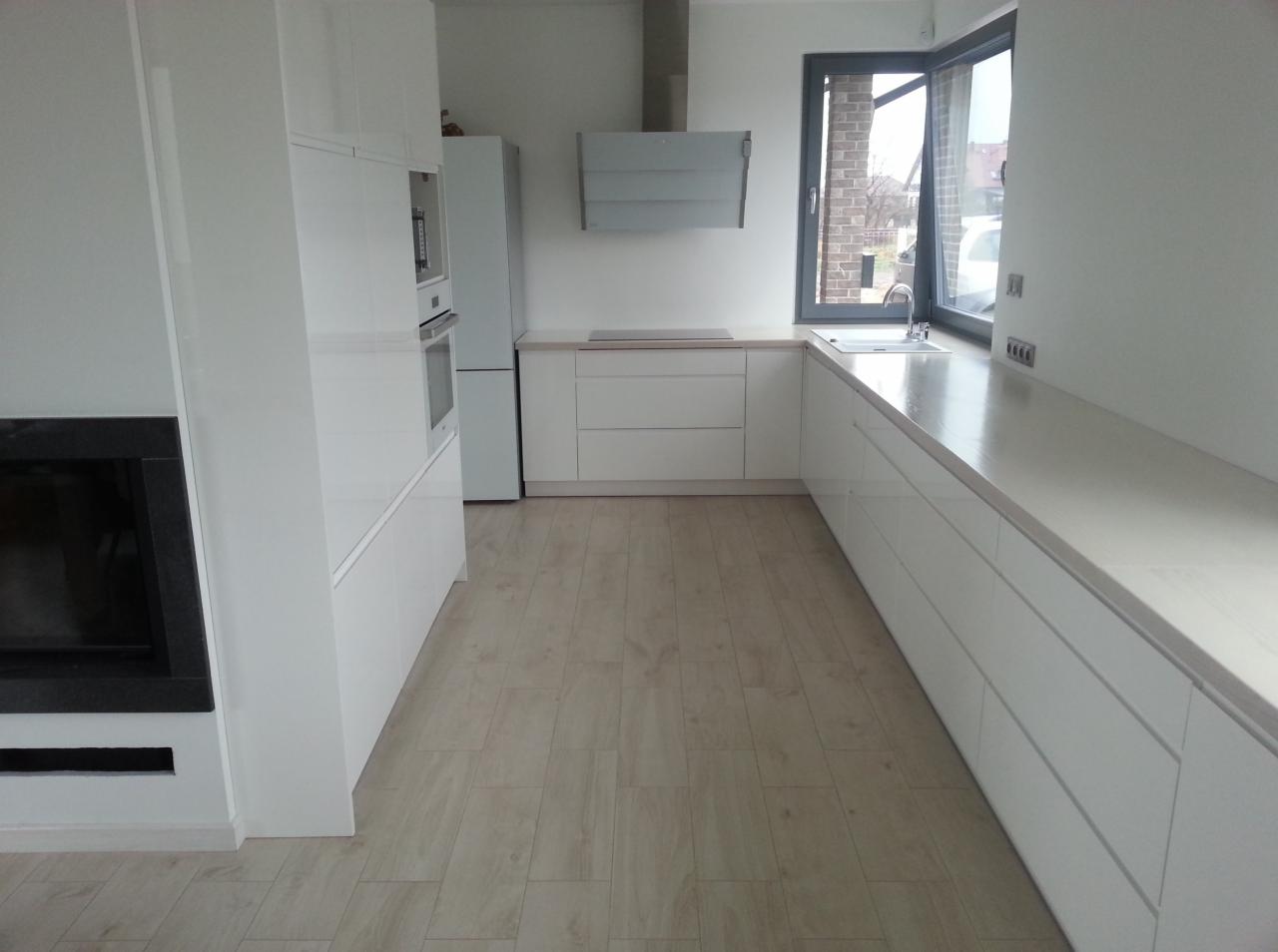 Biała kuchnia na wymiar z połyskiem, widok na okno i sprzęty AGD. Minimalistyczny design, jasne drewnopodobne panele podłogowe, czarny wkład kominkowy po lewej.