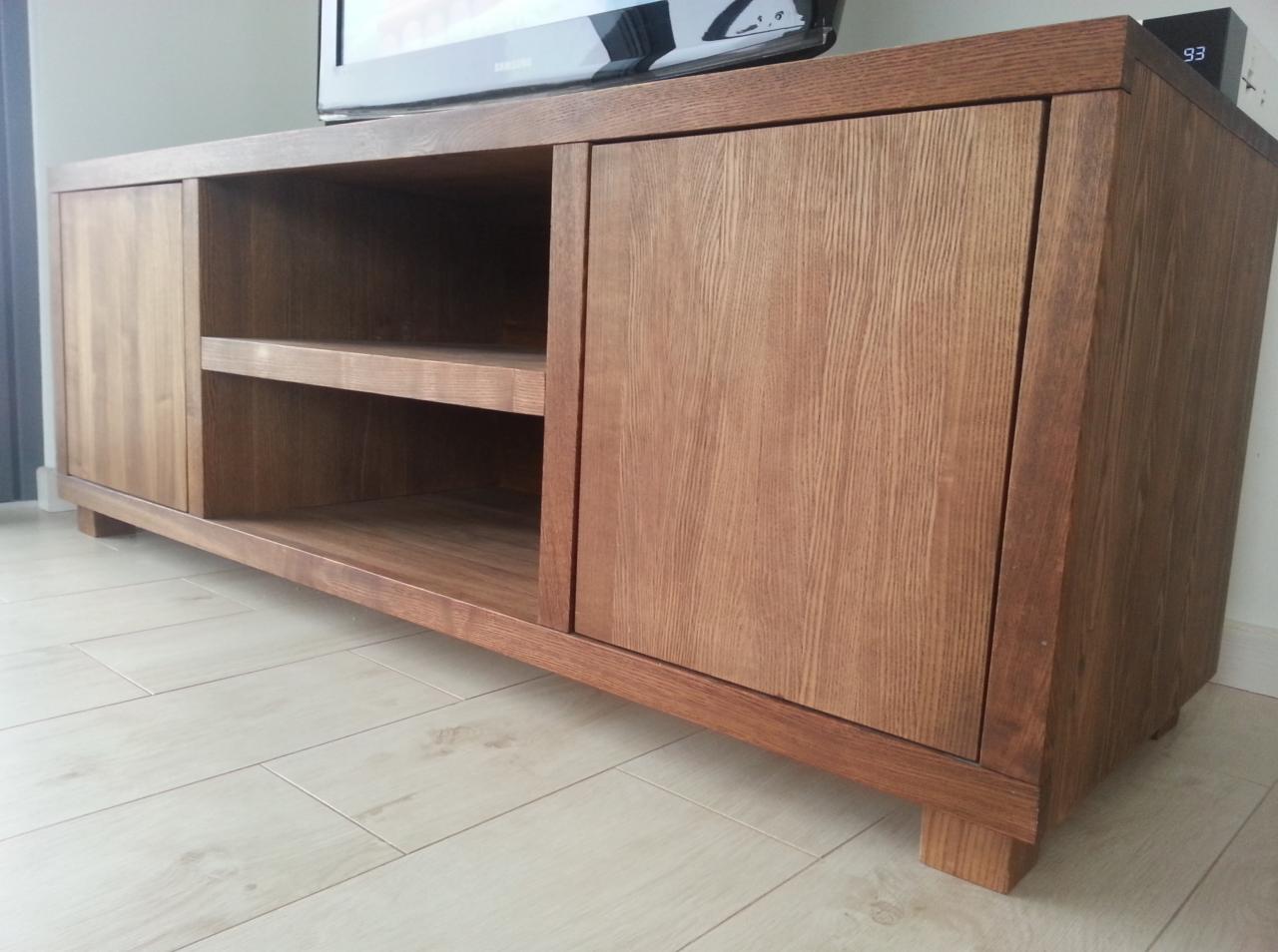 Drewniana szafka RTV z dwiema parami drzwi i otwartą półką, wykonana z dębowego drewna, stoi na jasnej podłodze. Na szafce stoi telewizor.