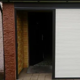 Biała, częściowo zasunięta osłona okienna na tle wejścia do budynku z brązową, teksturowaną elewacją. Widoczny fragment wnętrza i szara wycieraczka.