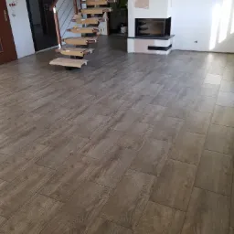 Ułożona podłoga z brązowych paneli imitujących drewno, widoczne schody drewniane w trakcie montażu z zabezpieczonymi kartonem stopniami, oraz kominek w trakcie wykończenia gładzią.