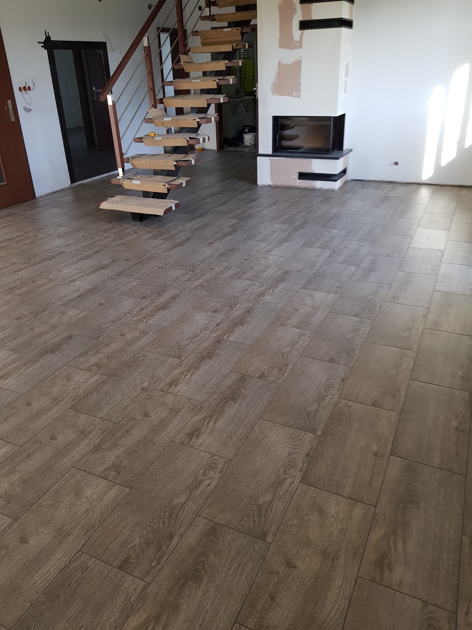 Ułożona podłoga z brązowych paneli imitujących drewno, widoczne schody drewniane w trakcie montażu z zabezpieczonymi kartonem stopniami, oraz kominek w trakcie wykończenia gładzią.