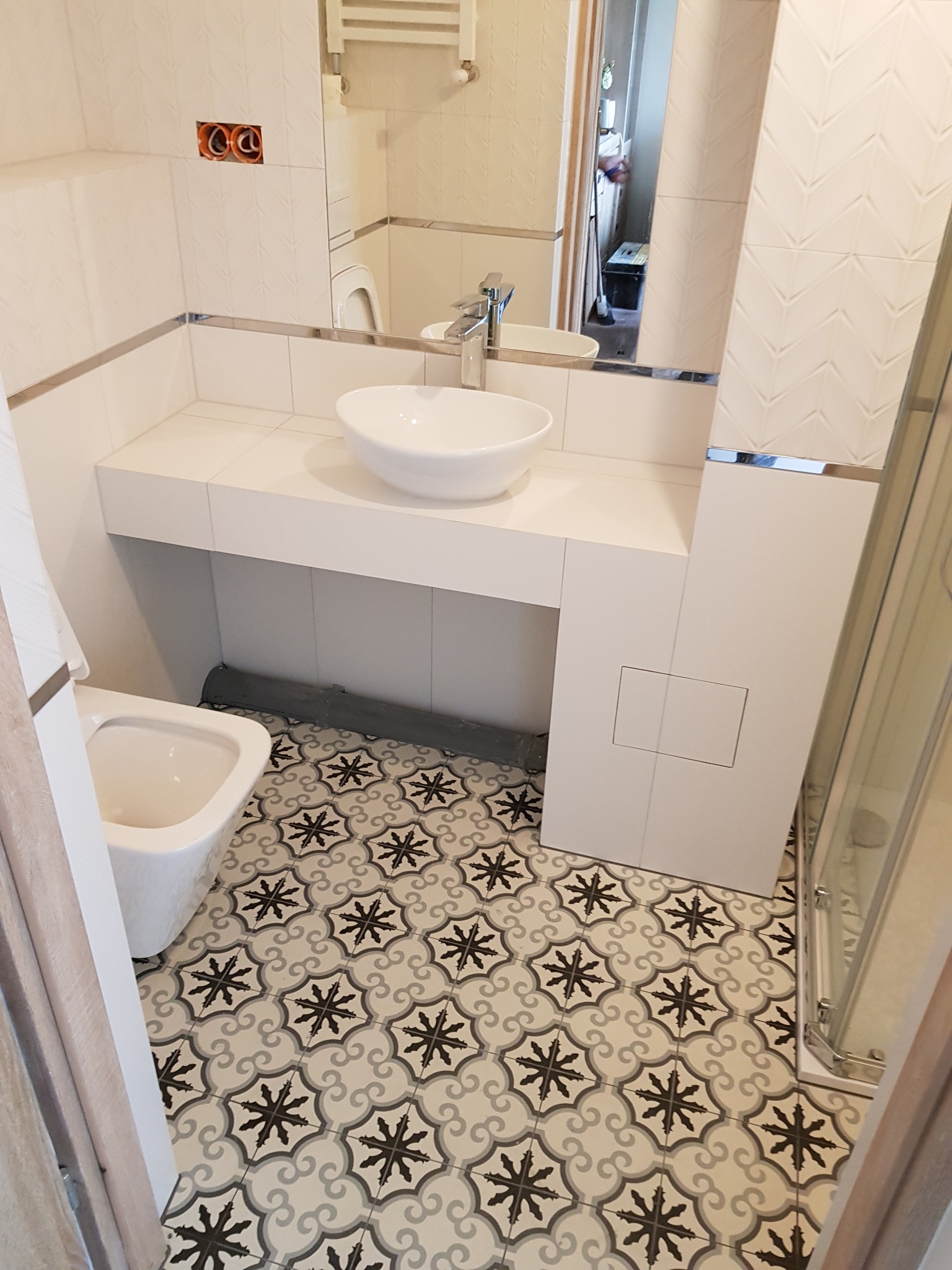 Wykończona łazienka z wzorzystą podłogą, białą armaturą i płytkami z delikatnym deseniem, widoczne niedokończone podłączenie elektryczne nad umywalką.