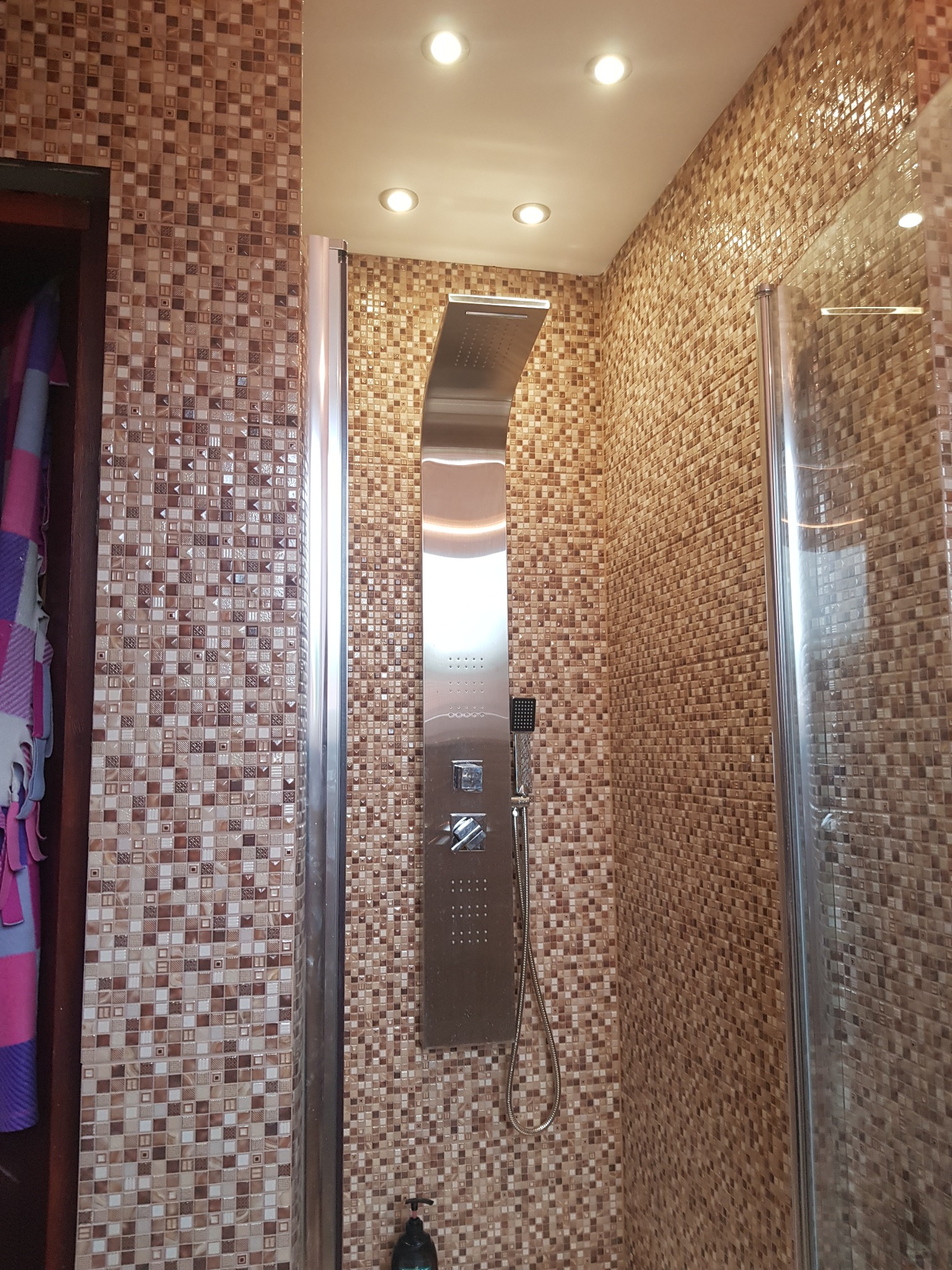 Wykończona kabina prysznicowa z mozaiką w odcieniach brązu i beżu, z panelem prysznicowym ze stali nierdzewnej i szklanymi drzwiami, widok z dołu.