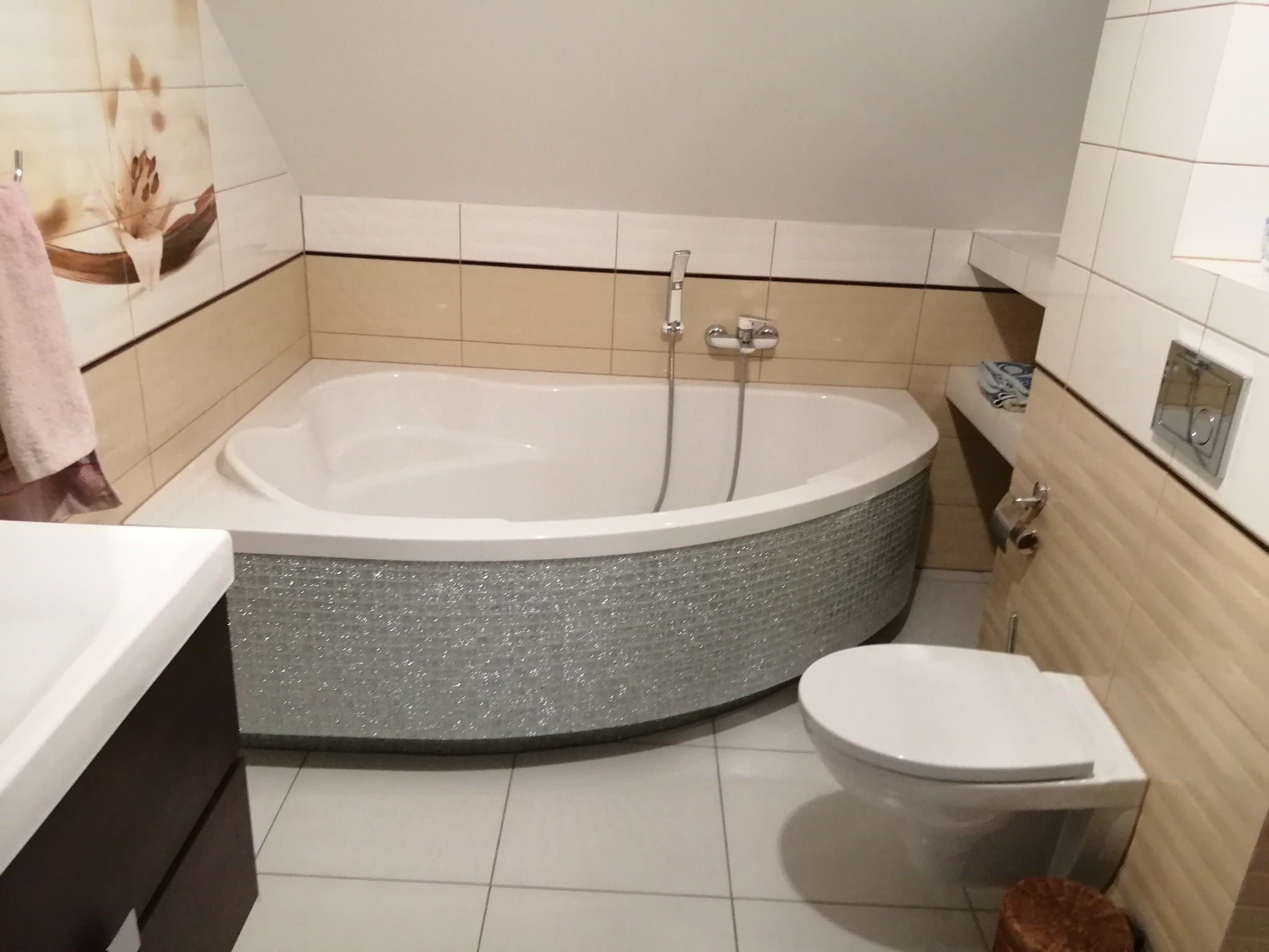 Wnętrze łazienki z narożną wanną obudowaną mozaiką, wiszącą toaletą i płytkami ściennymi w odcieniach beżu i bieli, z dekoracyjnym motywem kwiatowym na jednej ze ścian.