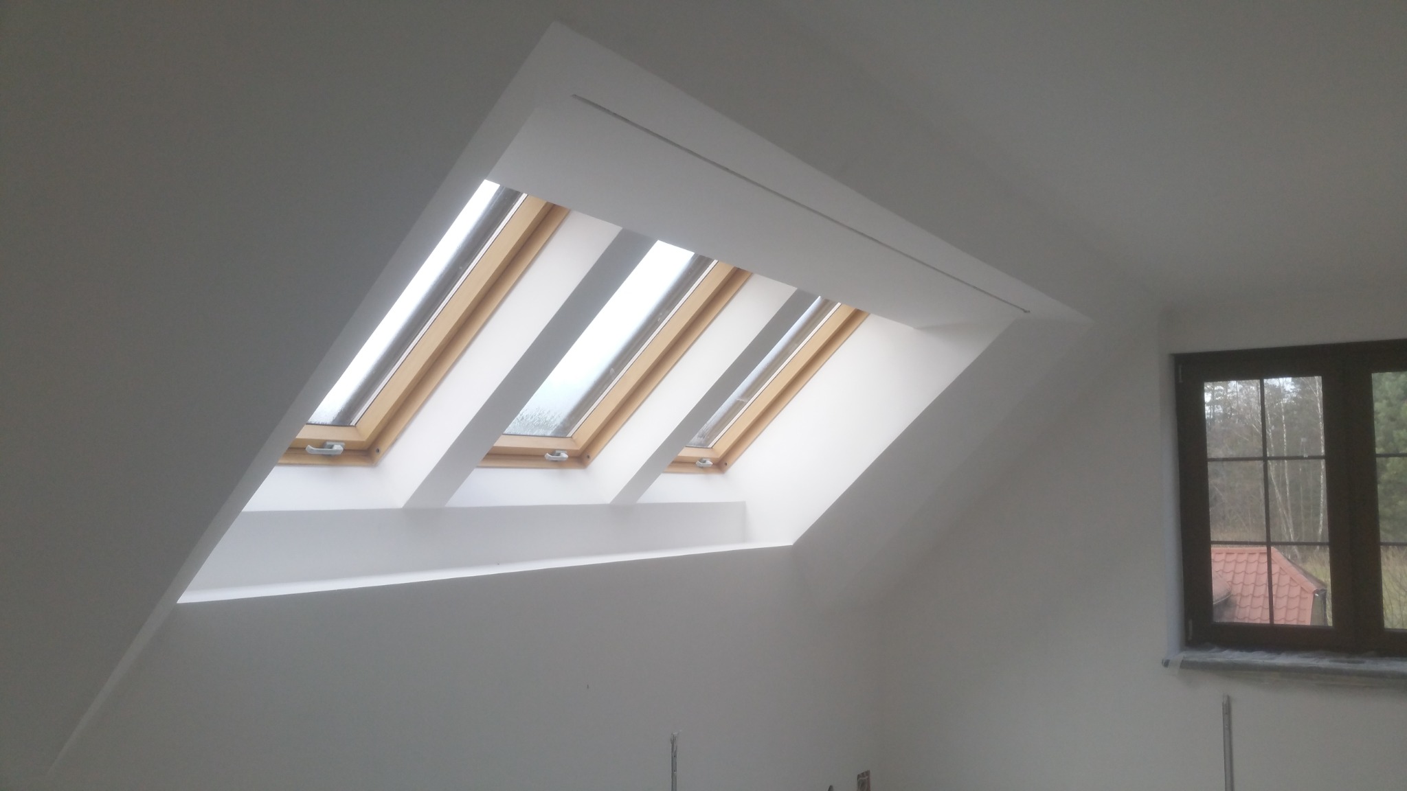 Poddasze po remoncie z trzema oknami dachowymi w drewnianej ramie, pomalowanymi na biało wnękami i ścianami, oraz dodatkowym oknem pionowym z widokiem na las.