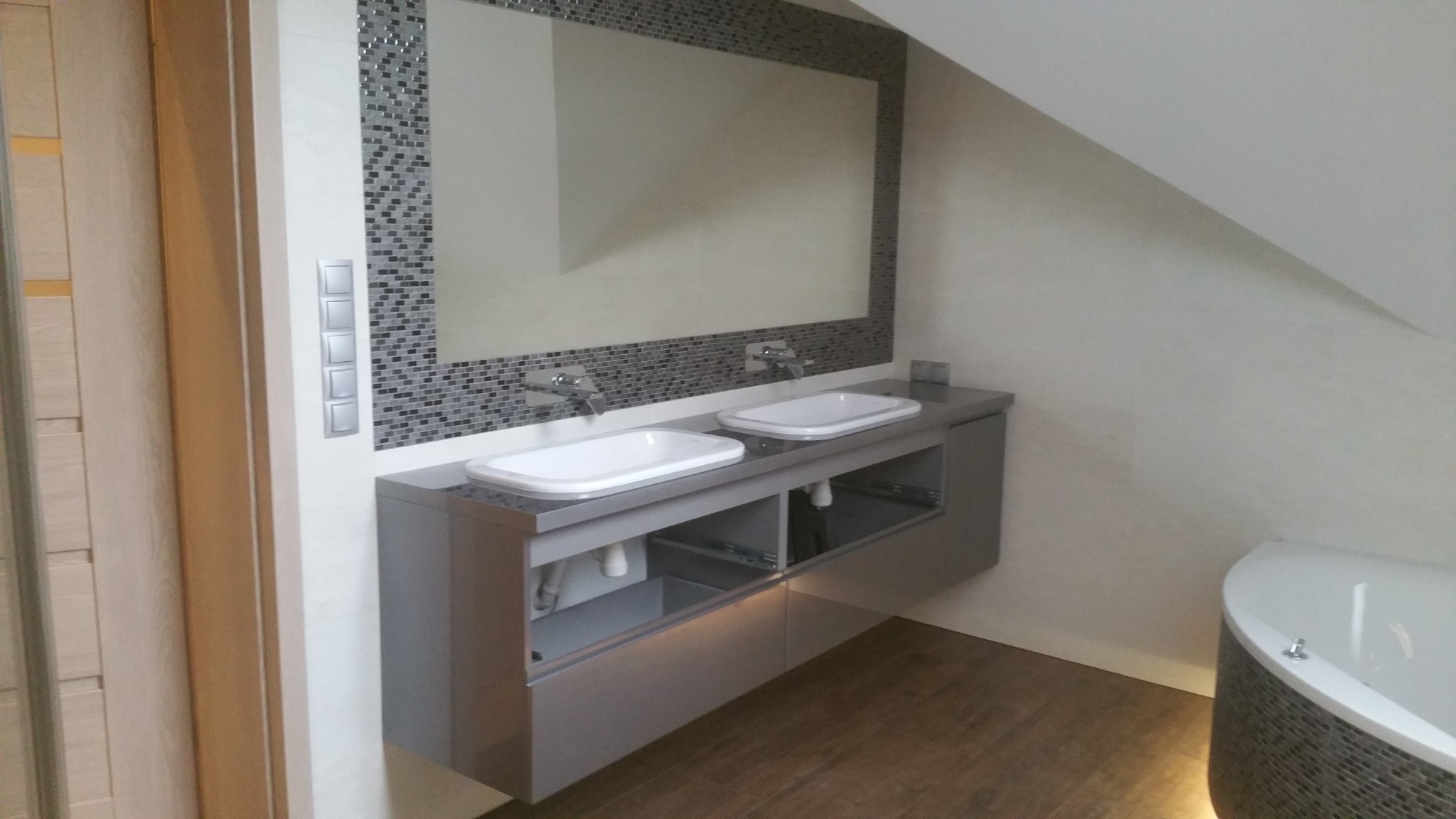 Nowoczesna łazienka na poddaszu z dwoma umywalkami nablatowymi na szarej szafce, lustro obramowane mozaiką, biała wanna z mozaiką na obudowie, drewnopodobna podłoga.