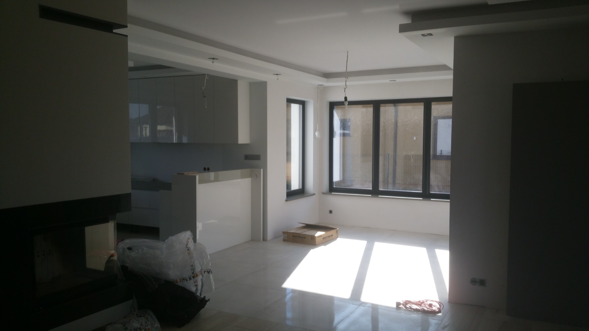 Jasne, przestronne wnętrze w trakcie wykończenia, widoczne świeżo położone, błyszczące płytki podłogowe, duże okna z czarnymi ramami, fragment zabudowy kuchennej i kominka, resztki materiałów...