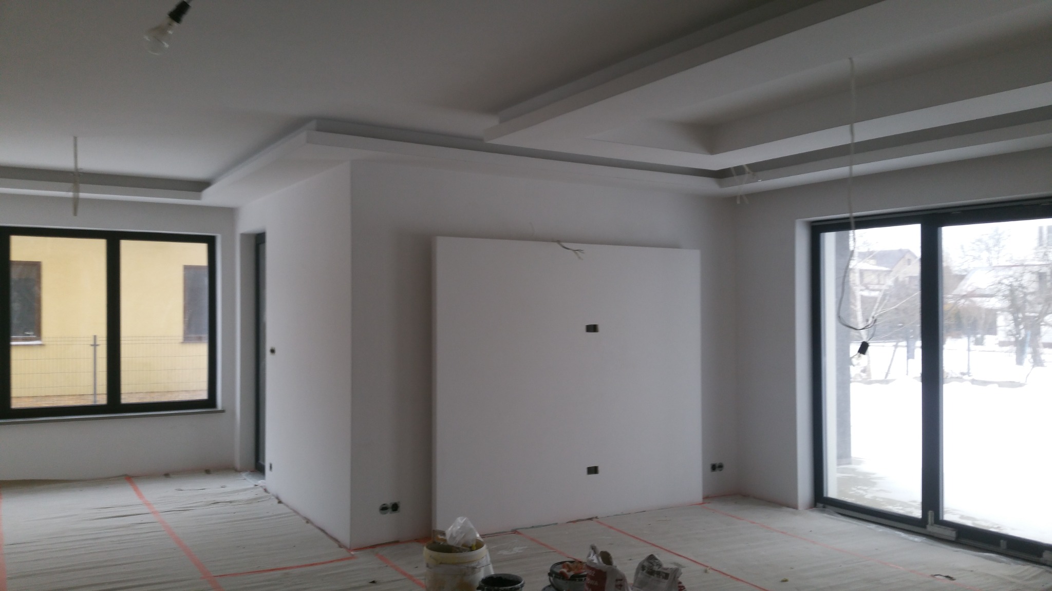 Wnętrze w trakcie wykańczania: białe ściany, sufit z dekoracyjnymi elementami, okna z czarnymi ramami, podłoga zabezpieczona folią malarską, widoczne puszki elektryczne i wiszące kable od oświetlenia.