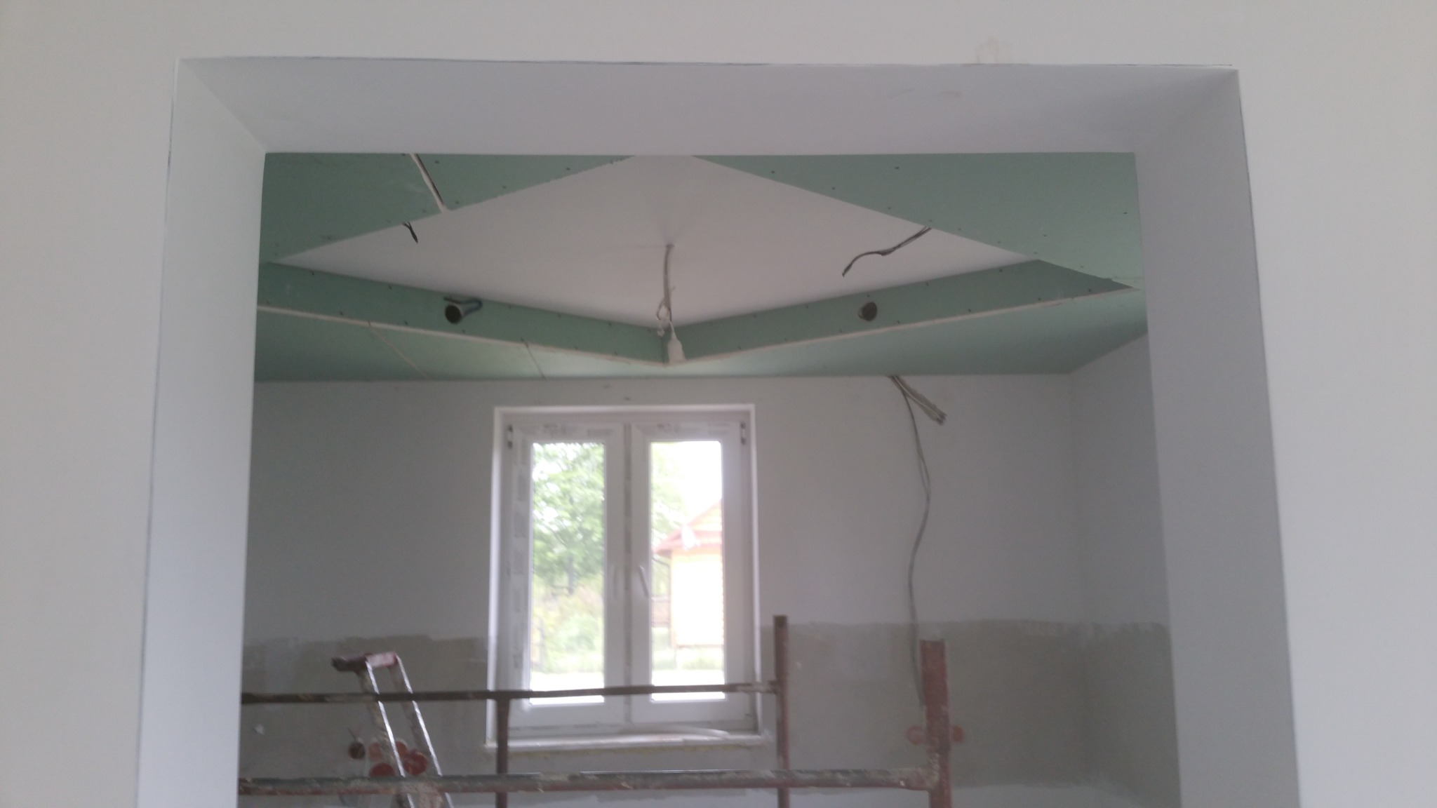 Wnętrze pokoju z widocznym fragmentem sufitu podwieszanego z płyty karton gips w trakcie montażu, widoczne kable i otwory na oświetlenie, w tle okno i rusztowanie.