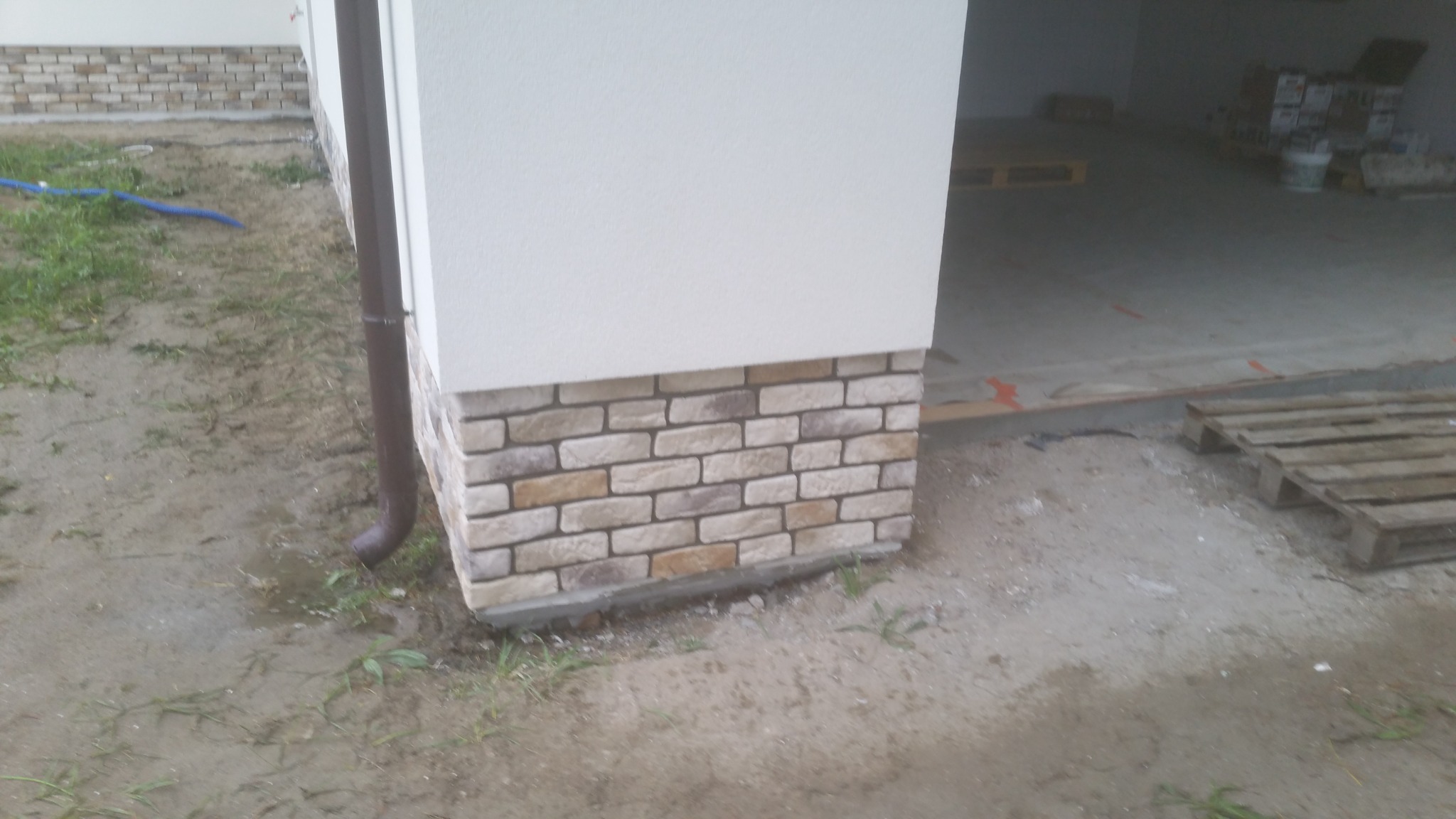 Wykończony fragment elewacji budynku z klinkierową okładziną, brązowa rynna spustowa, widoczne palety w otwartym garażu w tle.