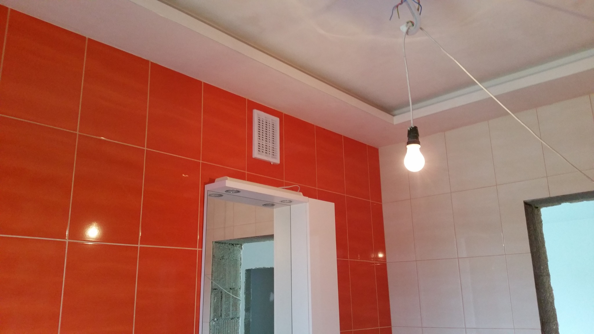 Pomarańczowe i białe płytki ceramiczne na ścianach łazienki, widoczny fragment lustra z oświetleniem, kratka wentylacyjna i prowizoryczne oświetlenie żarówką na kablu.