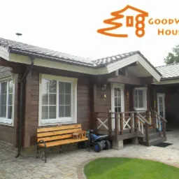 Parterowy dom z bali w brązowym kolorze, z białymi oknami i zadaszonym miejscem parkingowym. Na pierwszym planie ławka i mały rowerek.