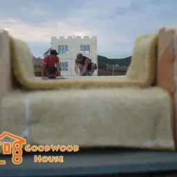 Praca na wysokości: trzech mężczyzn montuje izolację w drewnianej konstrukcji domu szkieletowego. W tle widać budynek w trakcie budowy i pagórkowaty krajobraz.