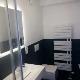 Nowoczesna łazienka z czarno-białymi płytkami, prostokątną umywalką z odpływem szczelinowym, grzejnikiem drabinkowym i oświetlonym lustrem z dotykowym włącznikiem.