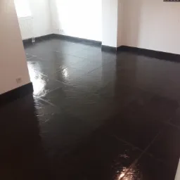 Błyszcząca, ciemna podłoga z płytek w pustym, jasnym pokoju z białymi ścianami i grzejnikiem, widoczny uchwyt do telewizora na ścianie.