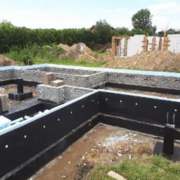 Budowa domu: Fundament z izolacją pionową i częściowo wymurowanymi ścianami z bloczków betonowych, widoczne szalunki i zbrojenia, w tle hałdy ziemi i konstrukcja domu w stanie surowym otwartym.